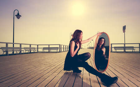 Solitude loneliness concept. Thoughtful young woman looks at the reflection in the mirror outdoors at sunsetの写真素材
