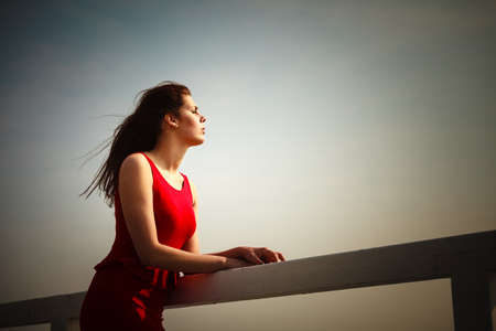 Holidays summer relaxation concept. Young thoughtful woman wearing red dress relaxing on pier outdoorの写真素材