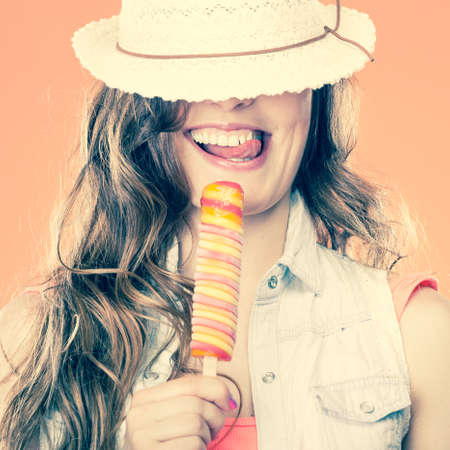Summer vacation happiness concept. Funny cheerful woman covering eyes with straw hat eating ice cream orange backgroundの写真素材
