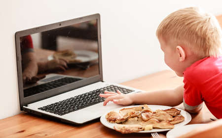 Children, technology and home concept  - little boy child eating meal while using laptop pc computer at home. Bad habitsの写真素材
