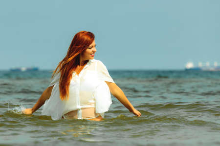 Vacation. Sensual girl wet cloth in water on the coast. Redhair woman having fun relaxing on the sea. Summertime.の写真素材