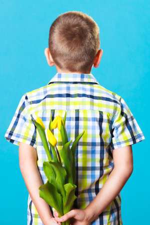 Holiday mother's day concept. Little boy has prepared surprise present for mum, flowers yellow tulips, holds it behind the back on blueの写真素材