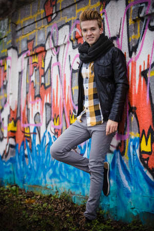 Full length of handsome trendy man outdoor in city setting, male model wearing black jacket scarf and checked shirt against colorful graffiti wallの写真素材