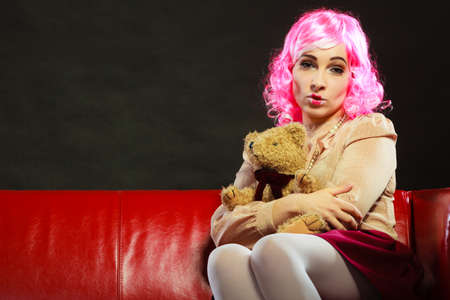 Mental disorder concept. Young childlike woman wearing like puppet doll sitting with teddy bear toy on red couch dark black backgroundの写真素材