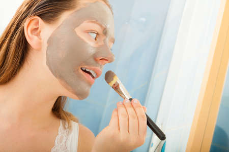 Beauty procedures skin care concept. Young woman applying facial gray mud clay mask to her face in...