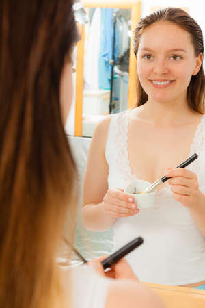 Beauty skin care cosmetics and health concept. Young woman applying facial mud clay mask to her face in bathroomの写真素材