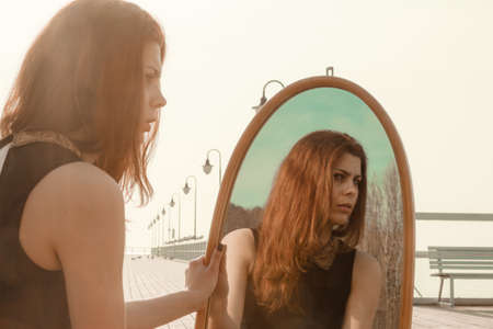 Solitude loneliness concept. Thoughtful young woman looks at the reflection in the mirror outdoors at sunsetの写真素材