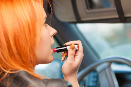 Concept of danger driving. Young woman driver red haired teenage girl painting her lips doing applying make up while driving the car.の写真素材