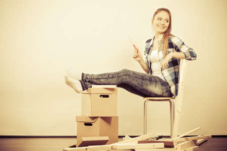 Woman moving into new apartment house assembling furniture with screwdriver. Young girl sitting on chair arranging interior and unpacking boxes.  の写真素材