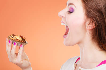 Bakery, sweet food and people concept. Woman face profile wide open mouth holds cake cupcake in hand orange backgroundの写真素材