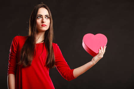 Charity and sharing concept. Love and feelings. Young pretty woman in red clothes holding heart box present gift on grey background in studio.の写真素材