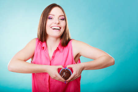 Sweet food sugar make us happy. Smiling woman holds chocolate muffin in hand making heart shape with fingers around cake blue backgroundの写真素材