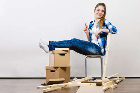 Woman moving into new apartment house assembling furniture with screwdriver. Young girl sitting on chair arranging interior and unpacking boxes.の写真素材