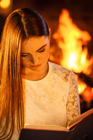 Woman reading book at fireplace. Young girl heating warming up and relaxing. Winter at home.