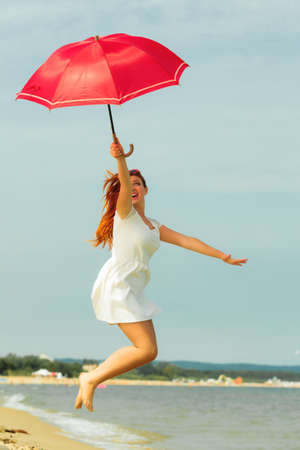 Holidays, vacation travel and freedom concept. Beautiful redhaired happy girl jumping with red umbrella on beach.の写真素材