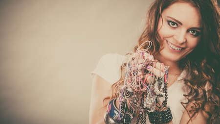Pretty young woman wearing bracelets and rings holding many plentiful of precious jewelry necklaces beads. Portrait of gorgeous fashion girl in studio on gray. の写真素材