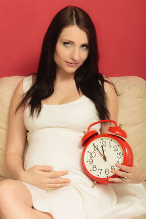 Pregnant woman holding red alarm clock waiting, sitting on sofa at homeの写真素材