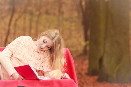 Happy smiling woman relaxing in fall park reading book. Young blonde girl resting laying on bench. Autumn lifestyle fun.の写真素材