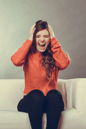 Angry furious outraged stressed young woman girl sitting on sofa couch at home. Negative emotions. Headache, pain.  の写真素材