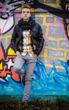 Full length of handsome trendy man outdoor in city setting, male model wearing black jacket scarf and checked shirt against colorful graffiti wallの写真素材