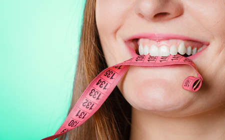 Diet and weight loss concept. Attractive woman holding pink measuring tape between teeth on blue background.の写真素材