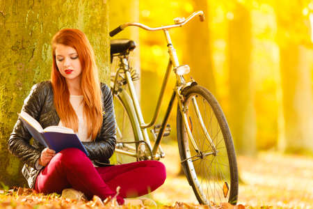Fall lifestyle concept. Redhaired young woman girl relaxing in autumn park reading book, sitting alone under tree. Sunny dayの写真素材
