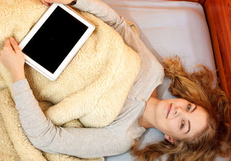 Young woman holding computer tablet with blank screen copyspace. Girl laying in bed under blanket....