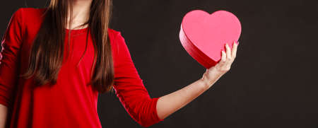 Valentines day, love, romance concept. Woman in red dress holding heart shaped gift box in hands dark gray background.の写真素材