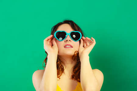 Summer fashion eyes protection concept. Closeup girl long curly hair in heart shaped sunglasses on green vivid color backgroundの写真素材