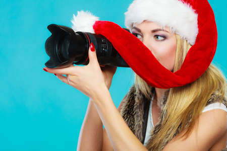 Photographer girl shooting images. Attractive blonde woman wearing santa claus helper hat taking photos with camera. Vivid blue backgroundの写真素材