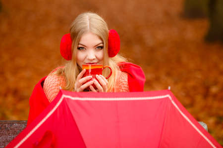 Happiness carefree and fall concept. Young happy woman relaxing in the autumn park on bench enjoying hot drink holding red mug with warm beverage. Orange leaves backgroundの写真素材
