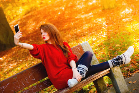 Technology internet and happiness concept. Woman happy girl taking self picture selfie with smartphone camera outdoors in autumn park on bench, golden leavesの写真素材