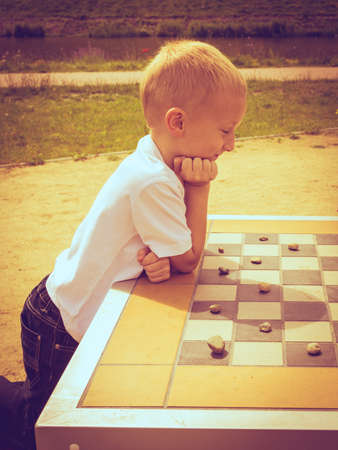 Draughts board game. Little boy clever child kid playing checkers thinking, outdoor in the park side...