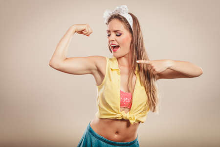 Happy strong pretty pin up girl with hairband bow showing off muscles. Young gorgeous retro woman in studio.の写真素材