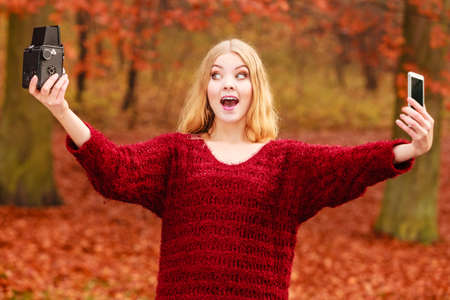 Pretty amazed woman in fall forest park taking selfie self photo with old vintage camera and smartphone. の写真素材