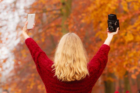 Pretty woman in fall forest park taking selfie self photo with old vintage camera and smartphone. Gorgeous young girl photographer. Autumn winter photography. Back rear view.の写真素材