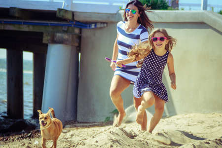 Happy carefree family with dog running on beach at sea coast. Little girl kid with mother having fun. Summer holidays vacation. Happiness.の写真素材