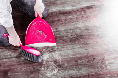 Cleanup housework concept. Closeup cleaning woman sweeping wooden floor with red small whisk broom and dustpan indoorの写真素材
