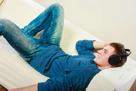 leisure and lifestyle concept. Young handsome man with headphones lying on couch listening music at homeの写真素材