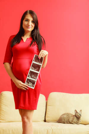 New life concept. Pregnancy, motherhood and happiness. Tummy of pregnant woman wearing elegant red dress, ultrasound scan in hand indoorの写真素材