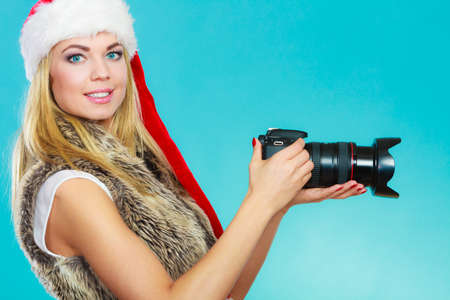 Photographer girl shooting images. Attractive blonde woman wearing santa claus helper hat taking photos with camera. Vivid blue backgroundの写真素材