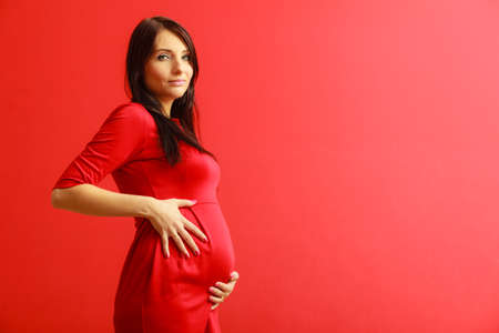 New life concept. Pregnancy, motherhood and happiness. tummy of pregnant woman wearing stylish elegant red dress indoorの写真素材
