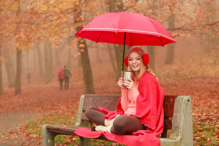 Woman with umbrella sitting on bench drinking hot cofee or tea relaxing in fall park. Young blonde girl resting outdoor. Autumn lifestyle fun.の写真素材