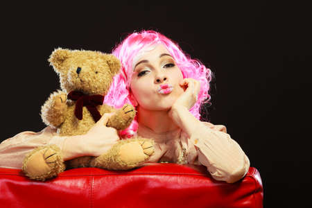 Mental disorder concept. Young childlike woman wearing like puppet doll sitting with teddy bear toy on red couch dark black backgroundの写真素材
