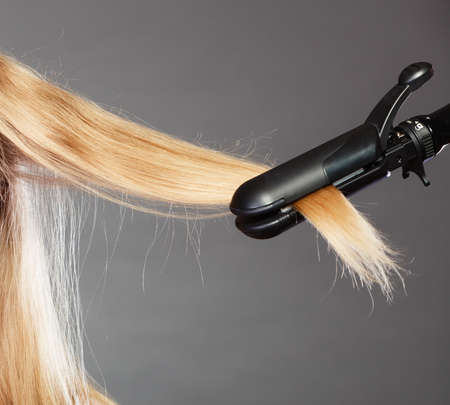Hairstyling. Closeup blonde woman long haired making hairstyle hairdo with electric hair iron straightener gray backgroundの写真素材