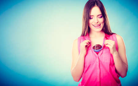 sweet food or sex for brighten moods. Smiling woman having fun holds chocolate muffin cake on her chest blue backgroundの写真素材
