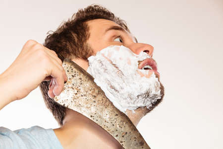 Scared cautious young man with shaving cream foam having fun with machete large knife. Handsome guy removing face beard hair. Skin care and hygiene humor.の写真素材
