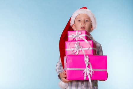Happy childhood, holiday christmas time.. Little boy child in red santa helper hat holding stack of pink presents gift boxes on blueの写真素材