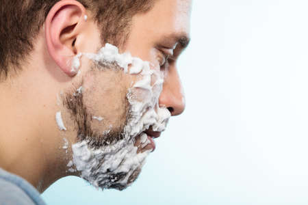 Health beauty and skin care concept. Closeup young bearded man with foam on face shaving on blue background.の写真素材