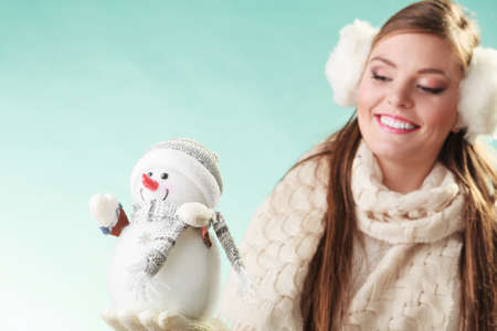 Smiling pretty cute woman holding little snowman. Attractive girl in earmuffs and white sweater pullover in studio on green. Winter fashion.の写真素材